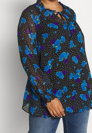 Woman wearing a sheer black blouse with blue and purple floral prints and white polka dots, paired with blue jeans.