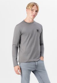 Cămașă cu mânecă lungă de culoare gri, realizată dintr-un material neted, având un guler rotund și o mică etichetă logo neagră pe partea stângă a pieptului.