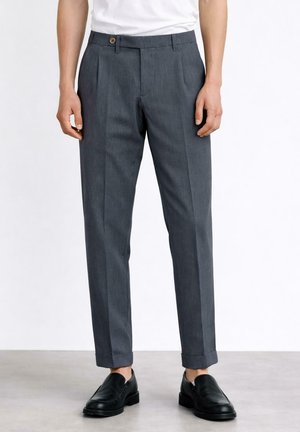 Mężczyzna noszący dopasowane ciemnoszare spodnie z mankietami i czarne skórzane wsuwane buty, stojący na jasnej podłodze.