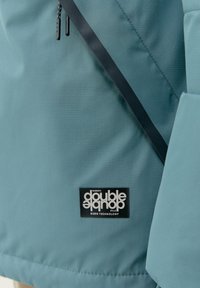 Zelena zunanja jakna s gladko teksturo, funkcionalnimi zippers in črnim logotipom z belim besedilom na spodnji levi strani.