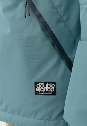 Tirkīza krāsas āra jaka ar gludu virsmu, funkcionālām rāvējslēdzēm un melnu logo uzšuvumu ar baltu tekstu apakšējā kreisajā stūrī.