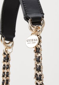 Schwarzes Lederarmband mit goldfarbenen Kettenakzenten und einem runden Anhänger, der "GUESS EST. 1981" zeigt und am Ende befestigt ist.