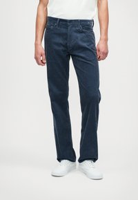 Sötétkék kordbársony nadrág egyenes szabással, függőleges bordázattal, öt zsebes kialakítással és fémből készült gombzárral. Fehér sportcipővel párosítva.