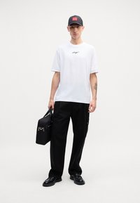 Hvid t-shirt med korte ærmer og sort skriftskriftlogo, parret med sorte brede bukser og sorte snørebåndssko. Holder en sort taske med sølvdetaljer.