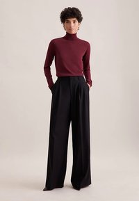 Bordeauxroter Rollkragenpullover kombiniert mit hoch taillierter, weit geschnittener schwarzer Hose. Glatter Stoff mit figurbetonter Silhouette und aufgesetzten Taschen vorne.