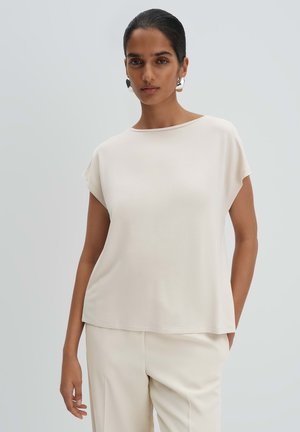 Donna con capelli scuri, indossa una maglietta a maniche corte color crema chiaro e pantaloni abbinati, con grandi orecchini d'argento, su uno sfondo chiaro e semplice.