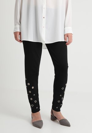 Schwarze Leggings mit dekorativen silbernen Ösen an den Seiten, kombiniert mit einer durchsichtigen weißen Bluse und grauen spitzen Ballerinas. Glatte Stofftextur.
