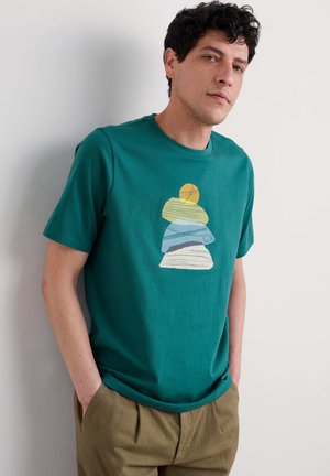 Camiseta de algodón en color verde azulado con un cuello redondo, que presenta un gráfico de piedras apiladas en colores amarillo, verde, azul y blanco. Fondo liso.
