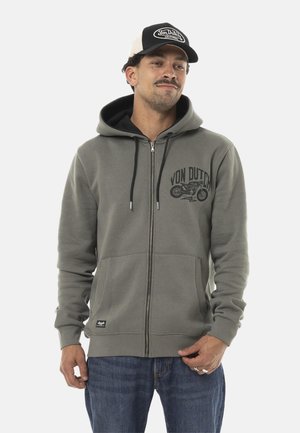 Homme portant un sweat à capuche gris zippé avec un motif de moto et le logo Von Dutch, jean bleu et casquette Von Dutch noire et blanche.