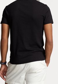 Muž oblečený v jednoduchém černém tričku s krátkým rukávem a bílých šortkách, stojící s jednou rukou v kapse, viděný ze zad.