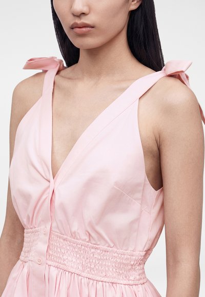 Γυναίκα που φορεί αμάνικο ανοιχτό ροζ φόρεμα με βαθύ V-λαιμόκοψη και μαζεμένη μέση, με τιράντες που δένονται σε φιόγκο στους ώμους.