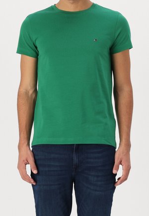 Mann trägt ein schlichtes grünes kurzärmliges T-Shirt mit kleinem Logo auf der Brust und dunkelblaue Jeans, steht vor einem hellen Hintergrund.