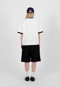 Personne aux longs cheveux blonds portant un t-shirt blanc à bordure noire, un short noir, des chaussettes blanches, des chaussures noires et une casquette noire, vue de dos.