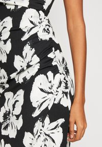 Detailní záběr černých šatů s velkými bílými květinovými potisky, zobrazující část trupu a ruku visící vedle těla.