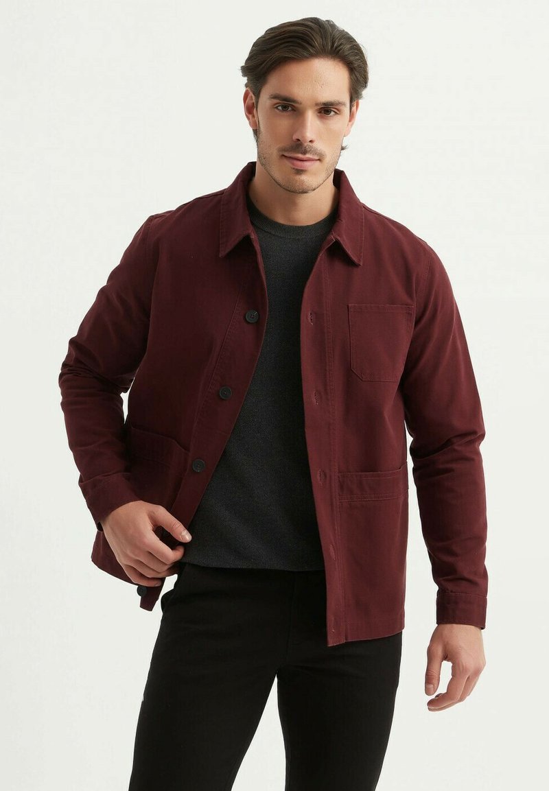 Veste boutonnière bordeaux en coton. Dotée d'un col, de poches avant et d'un tissu texturé. Portée par-dessus un pull sombre et un pantalon noir.