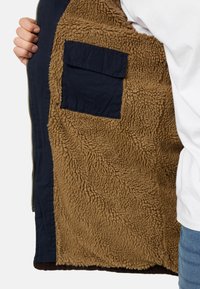 Chaqueta azul marino con forro de piel sintética marrón, que presenta un bolsillo tipo parche y un cierre de cremallera. Interior texturizado y dobladillo acanalado.