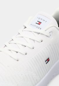 Weiße Sneaker mit strukturiertem Mesh-Obermaterial, flachen Schnürsenkeln und einem Logo-Label. Verfügt über dezente Streifenakzente in Rot, Weiß und Blau an der Seite.