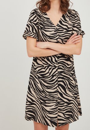 Kobieta w sukience z krótkim rękawem i dekoltem w serek, w czarno-beżowy wzór zebry, stojąca z założonymi rękami na tle jednolitego koloru.
