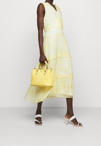 Gul, lager-på-lager maxi klänning med blommönster; en vadderad gul handväska med dubbla handtag; vita sandaler med remmar.