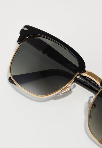 Sunglasses with black plastic frames and gold metal accents. Dark gray lenses and transparent nose pads. Slightly oversized square shape.