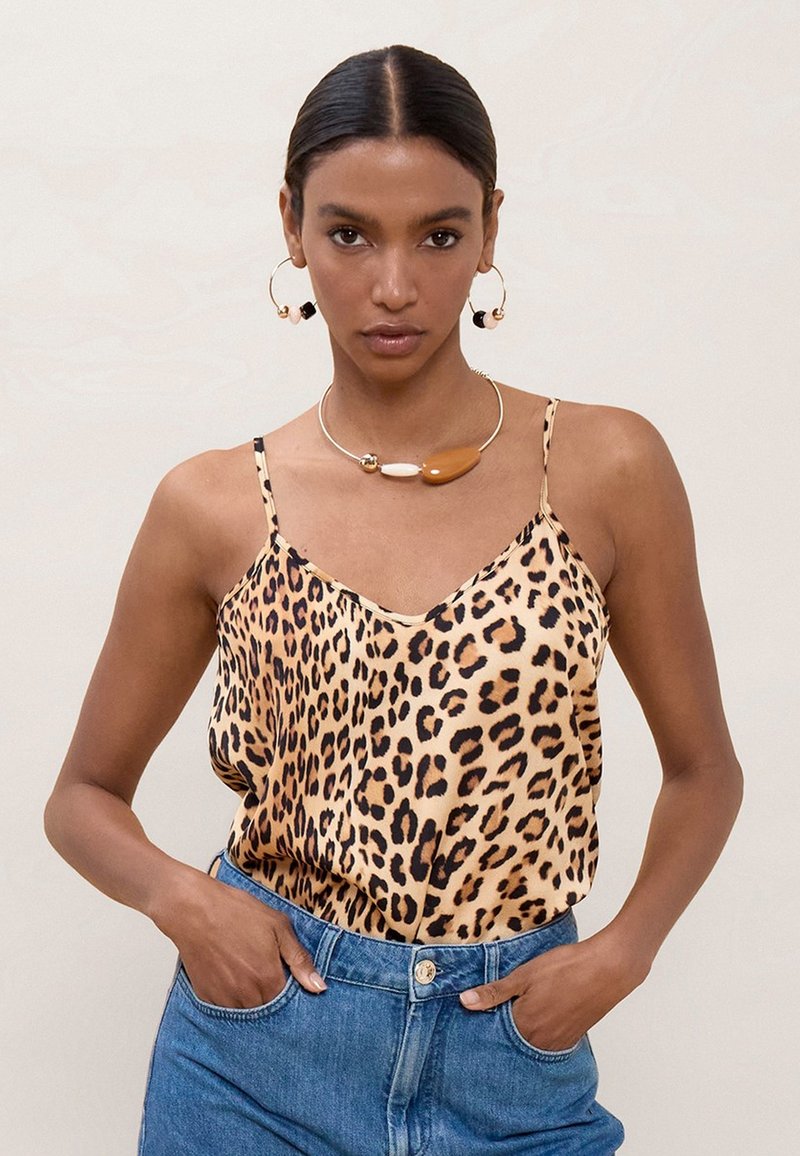 Žena v tielku s leopardím vzorom, modré džínsy, veľké kruhové náušnice a hrubý dvojfarebný náhrdelník, ruky v vreckách.