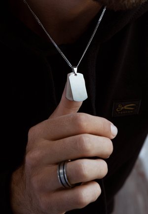 Zilveren dogtag-hanger aan een ketting, met een geborstelde afwerking en afgeronde randen, vastgehouden door een hand met een donker gekleurd ring.