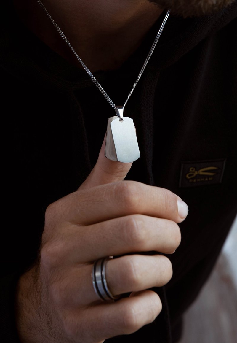 Silberner Hundemarke-Anhänger an einer Kette, mit gebürsteter Oberfläche und abgerundeten Kanten, gehalten von einer Hand mit einem dunkel gefärbten Ring.