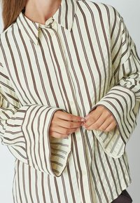 Chemise crème à boutons avec des rayures verticales marron, dotée de manches larges et d'un col classique. Tissu léger et texturé.