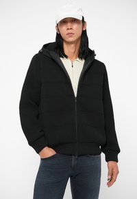 Crna podstavljena jakna s rebrastom teksturom, kapuljačom i zatvaranjem na zip, nosi se preko svijetle košulje, u kombinaciji s tamnim trapericama. Ima opušteni kroj.