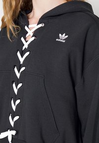 Felpa nera con cappuccio e dettagli in pizzo bianco, dotata di tasca a marsupio e piccolo logo Adidas bianco sul petto sinistro.