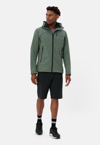 Grüne Softshelljacke mit Kapuze, Frontreißverschluss und zwei Seitentaschen, kombiniert mit schwarzen Shorts und blauen Wanderschuhen, stehend vor einem einfarbigen Hintergrund.