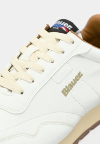 Baskets en cuir blanc avec des accents beiges, dotées de lacets plats beiges, d'un logo doré sur le côté et d'une semelle extérieure en caoutchouc.