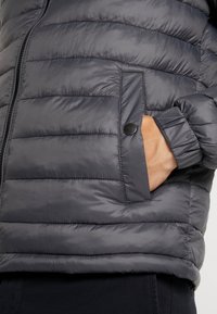 Gewatteerd jack in donkergrijs met horizontale stiksels, voorzien van een zak met drukknopen en elastische manchetten. Gladde, glanzende textuur.