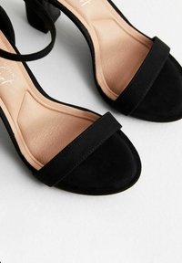 Sandali con tacco alto in suede nero, dotati di una larga fascia anteriore e di una soletta beige liscia. Presentano un tacco a blocco e un design minimalista.