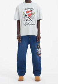 Szara koszulka z krótkim rękawem z grafiką czaszki i serca, zestawiona z luźnymi niebieskimi dżinsami z kolorową haftowaną dekoracją na nogawce. Brązowe buty.