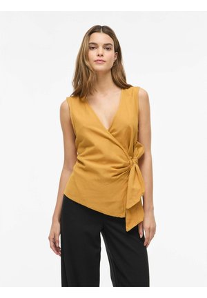 Femme portant un haut cache-cœur sans manches moutarde avec un lien sur le côté, associé à un pantalon large noir, debout sur un fond blanc uni.