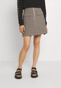 Geruite mini-jurk in zwart, rood en groen met een rits aan de voorkant en twee opgestikte zakken; gecombineerd met zwarte sandalen.