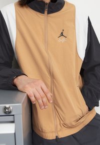 Ruskea ja musta vetoketjullinen takki valkoisilla yksityiskohdilla; siinä on Jordan-logo ja joustavat hihansuut; valmistettu kevyestä, sileästä kankaasta.