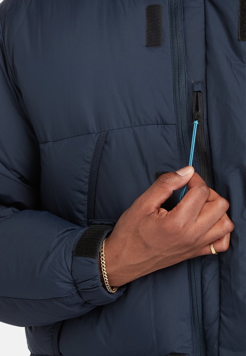 Ręka z złotą bransoletką i pierścionkiem ciągnie niebieski suwak na ciemnoniebieskiej ocieplanej kurtce z rzepami na rękawie.