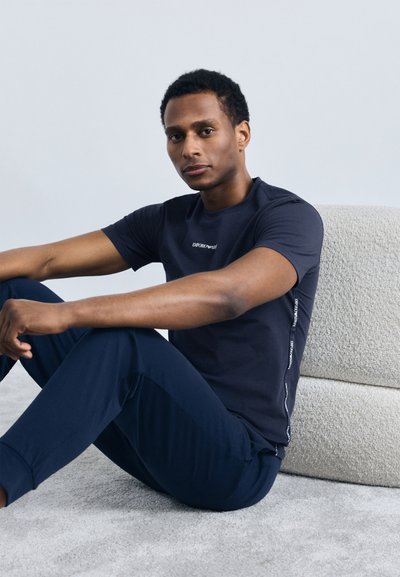 Sēdošs vīrietis, tērpies tumši zilā Emporio Armani T-kreklā un biksēs, atpūšas pret teksturētu gaišas krāsas spilvenu sēdekli.