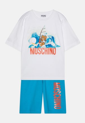 Biela bavlnená tričko s grafiku medveďa, s dizajnom surfovacích dosiek a vlny. Modré šortky s nápisom "MOSCHINO" v oranžovom texte na boku.