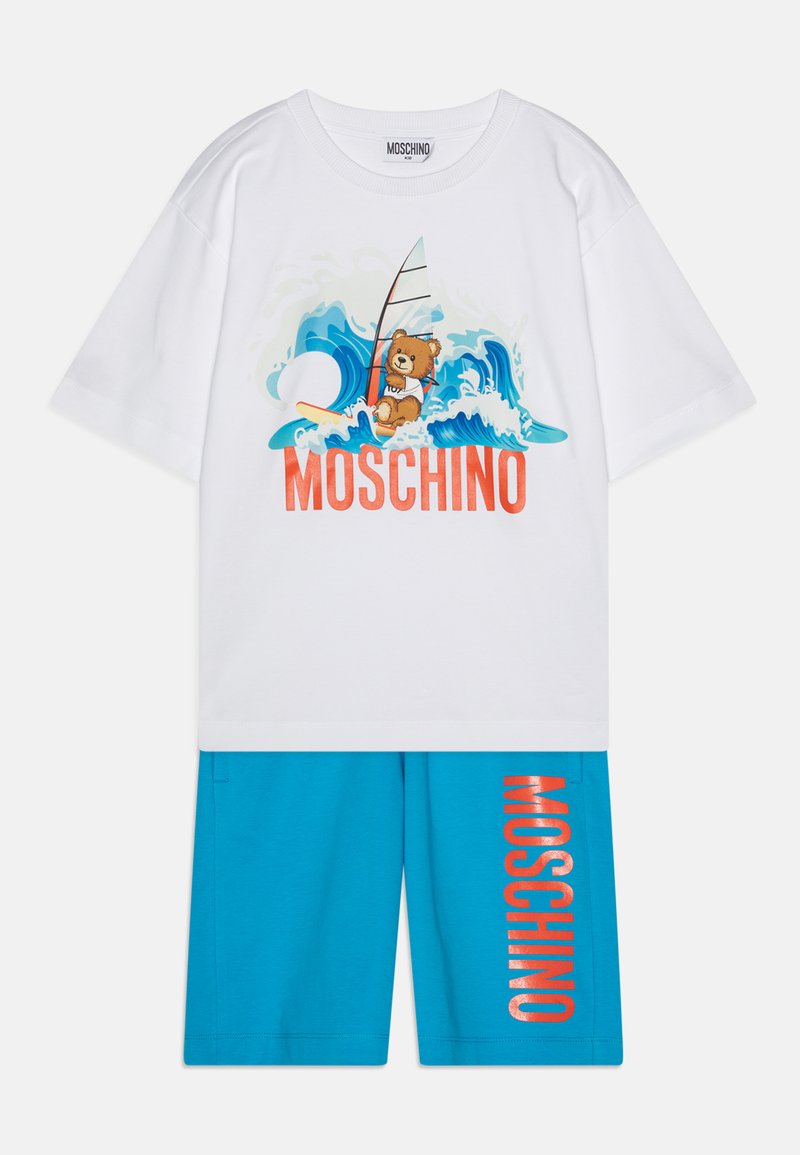 Bela bombažna majica z grafiko medveda, s surf desko in valovnim vzorcem. Modri šortsi z napisom "MOSCHINO" v oranžni barvi na strani.