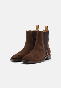 Bottes chevilles en daim marron avec des panneaux élastiques sur les côtés, des bouts pointus et des languettes de traction. Elles possèdent un talon bas empilé et un accent en cuir lisse.