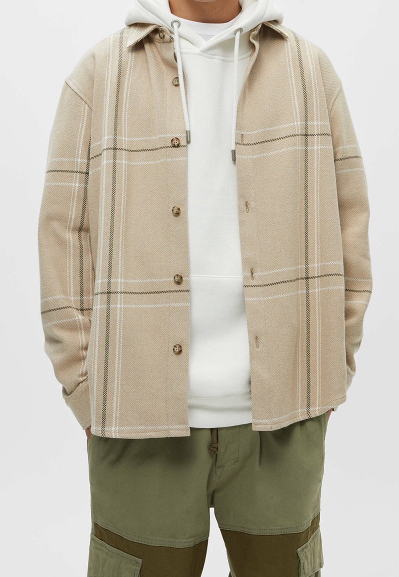 Personne portant une chemise beige à carreaux boutonnée par-dessus un sweat à capuche blanc avec cordons et un pantalon cargo vert olive avec poches.