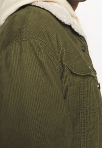 Giubbotto in velluto a coste verde oliva con colletto in pelliccia sintetica panna, dotato di chiusura con bottoni e tasche laterali, che presenta una texture a coste.