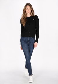 Musta ribattu neuletakki pitkillä hihoilla, pyöreällä kaula-aukolla ja etunapeilla. Yhdistetty sinisiin slim-fit farkkuihin ja valkoisiin tennareihin.