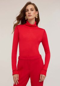 Top roșu cu guler înalt, confecționat dintr-un material moale, având mâneci lungi cu manșete cordonate. Asortat cu pantaloni roșii cu talie înaltă.