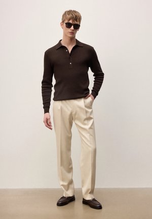 Mladý muž s čiernymi slnečnými okuliarmi, tmavohnedou polokošeľou s dlhým rukávom, béžovými nohavicami a tmavohnedými topánkami stojí pred jednofarebným pozadím.