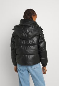 Persona con cabello trenzado que lleva una chaqueta de plumas negra con capucha y jeans de color azul claro, de pie contra un fondo claro y liso.
