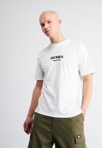 Camiseta blanca de algodón con el logotipo "DICKIES" en negro y el texto "SINCE 1922". El modelo lleva pantalones cargo en verde olivo, mostrando un ajuste relajado.